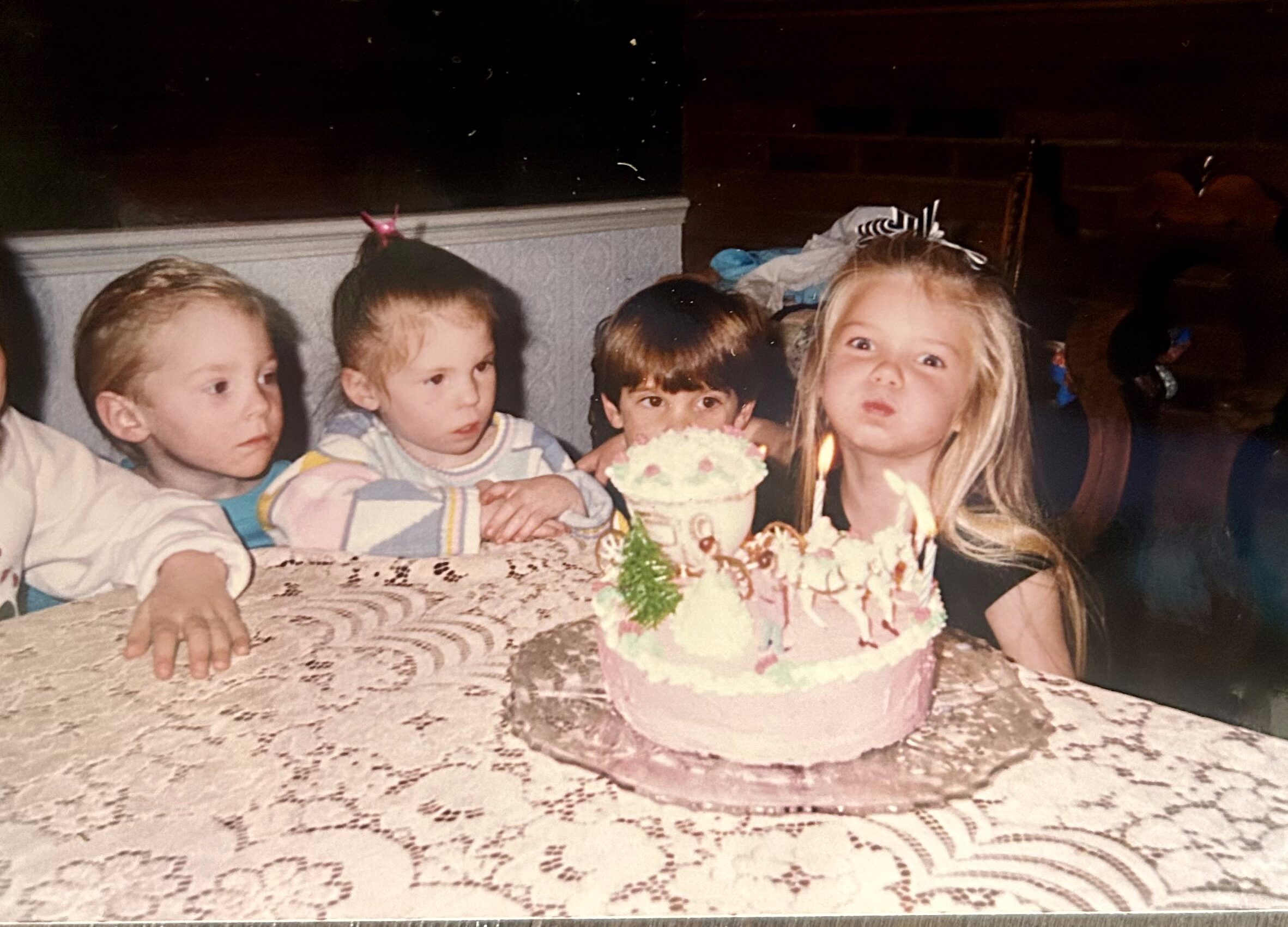 blonde little girl blowing out birthday candles while friends sit around the table watching